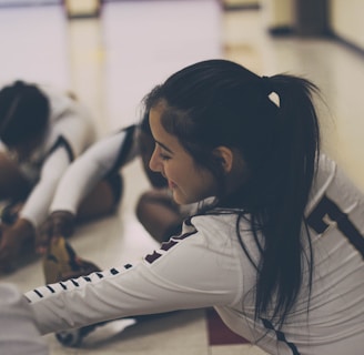A group of athletes wearing sports uniforms is seated on the floor in a gymnasium, engaging in stretching exercises. The focus is on one athlete with a ponytail, wearing a jersey with the number seven, smiling as she stretches her legs. The lighting is soft, creating a relaxed atmosphere.