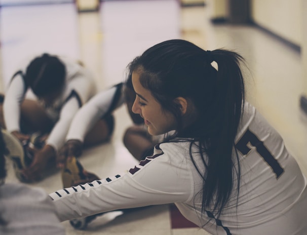 A group of athletes wearing sports uniforms is seated on the floor in a gymnasium, engaging in stretching exercises. The focus is on one athlete with a ponytail, wearing a jersey with the number seven, smiling as she stretches her legs. The lighting is soft, creating a relaxed atmosphere.