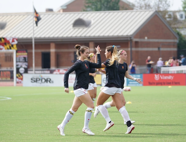 Three women in black athletic shirts and white shorts are on a grassy field, engaged in some activity that appears to be related to sports practice or warm-up. They are in an open area with a building with large windows in the background. There are also a few people visible near the building. The field is marked for a sporting event, and a few pieces of sports equipment can be seen, including a soccer ball and some training cones.