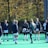 A group of people, primarily young women, wearing athletic uniforms and jackets are standing in a circle on a green sports field. One person is in the center of the circle, appearing to balance a ball on their knee. The background features trees with autumn foliage.