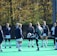 A group of people, primarily young women, wearing athletic uniforms and jackets are standing in a circle on a green sports field. One person is in the center of the circle, appearing to balance a ball on their knee. The background features trees with autumn foliage.