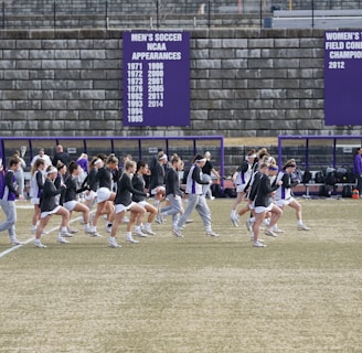 A group of athletes in sports attire are exercising or warming up on a grassy field. They are in a formation, appearing to be mid-stride, and the background shows a series of purple banners with sports achievements listed. The area is surrounded by a stone wall with benches and equipment visible.