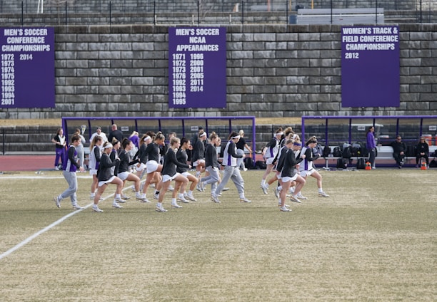 A group of athletes in sports attire are exercising or warming up on a grassy field. They are in a formation, appearing to be mid-stride, and the background shows a series of purple banners with sports achievements listed. The area is surrounded by a stone wall with benches and equipment visible.