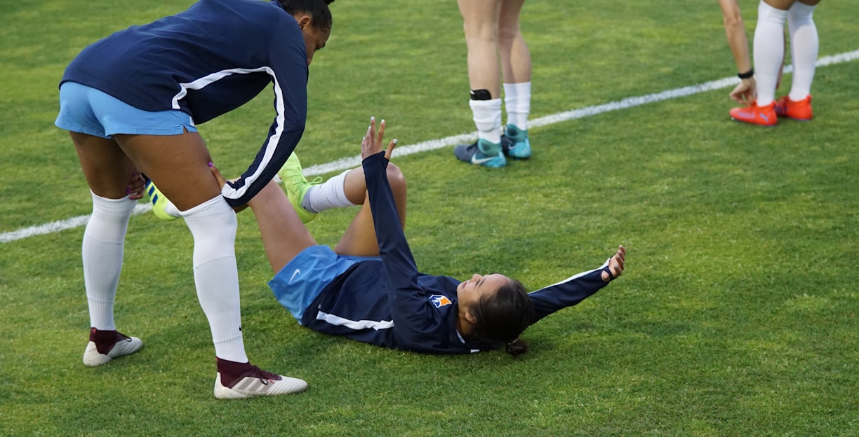 A group of athletes in sportswear are on a green soccer field. Two individuals are interacting closely; one helps the other who is lying on the grass. Nearby, other athletes are stretching.