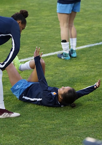 A group of athletes in sportswear are on a green soccer field. Two individuals are interacting closely; one helps the other who is lying on the grass. Nearby, other athletes are stretching.