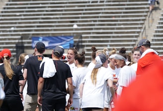A group of athletes in white shirts with black and red accents are gathered together, engaged in conversation and smiling. They appear to be on a sports field with empty bleachers in the background. The setting is outdoors, and it seems sunny. Some individuals are wearing caps or headbands.
