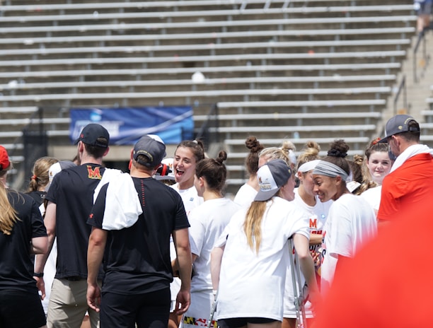 A group of athletes in white shirts with black and red accents are gathered together, engaged in conversation and smiling. They appear to be on a sports field with empty bleachers in the background. The setting is outdoors, and it seems sunny. Some individuals are wearing caps or headbands.