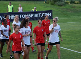 A group of women in sports attire are gathered on a grassy field, casually interacting and smiling. They are wearing shirts with 'Washington Spirit' written on them, suggesting they are part of a soccer team. A banner is visible in the background displaying the same name, reinforcing the team atmosphere. The image captures a moment of camaraderie and light-heartedness among the players.