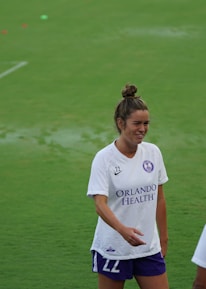 A woman wearing a white sports jersey with 'Orlando Health' written on it stands on a grassy field, smiling and engaging with another person who is partially visible. She has her hair tied up and is wearing shorts. The field is well-maintained and appears to be an athletic or sports setting.