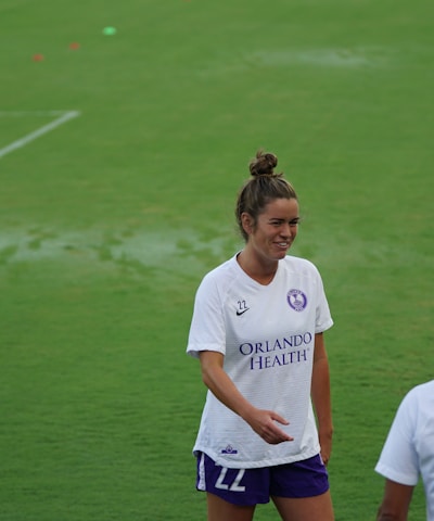 A woman wearing a white sports jersey with 'Orlando Health' written on it stands on a grassy field, smiling and engaging with another person who is partially visible. She has her hair tied up and is wearing shorts. The field is well-maintained and appears to be an athletic or sports setting.