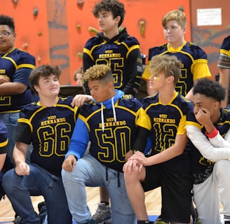 A group of young athletes wearing blue and yellow jerseys are gathered in a gym with climbing walls in the background. The jerseys have numbers and the words 'West Hernando' on them. Some are standing, while others are kneeling, showing camaraderie and team spirit.