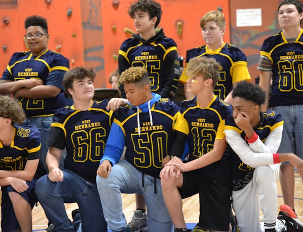 A group of young athletes wearing blue and yellow jerseys are gathered in a gym with climbing walls in the background. The jerseys have numbers and the words 'West Hernando' on them. Some are standing, while others are kneeling, showing camaraderie and team spirit.