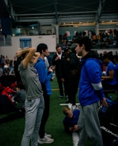 People engage in conversation and activities in an indoor sports facility. A woman in gray athletic wear stretches her arms above her head while talking to a man in a blue jacket. Other people are seen interacting in the background, some are seated on the ground, and the atmosphere is lively with many individuals gathered.