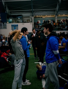 People engage in conversation and activities in an indoor sports facility. A woman in gray athletic wear stretches her arms above her head while talking to a man in a blue jacket. Other people are seen interacting in the background, some are seated on the ground, and the atmosphere is lively with many individuals gathered.
