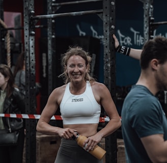 A woman in athletic wear is standing in a gym environment, smiling and holding a water bottle. She appears to be in front of gym equipment with other people around. The atmosphere looks energetic and focused.