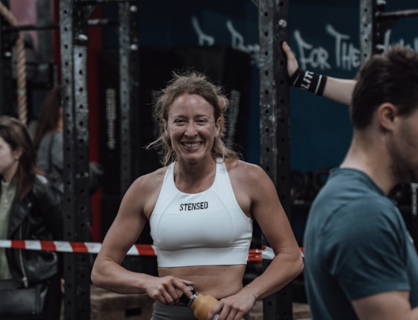 A woman in athletic wear is standing in a gym environment, smiling and holding a water bottle. She appears to be in front of gym equipment with other people around. The atmosphere looks energetic and focused.